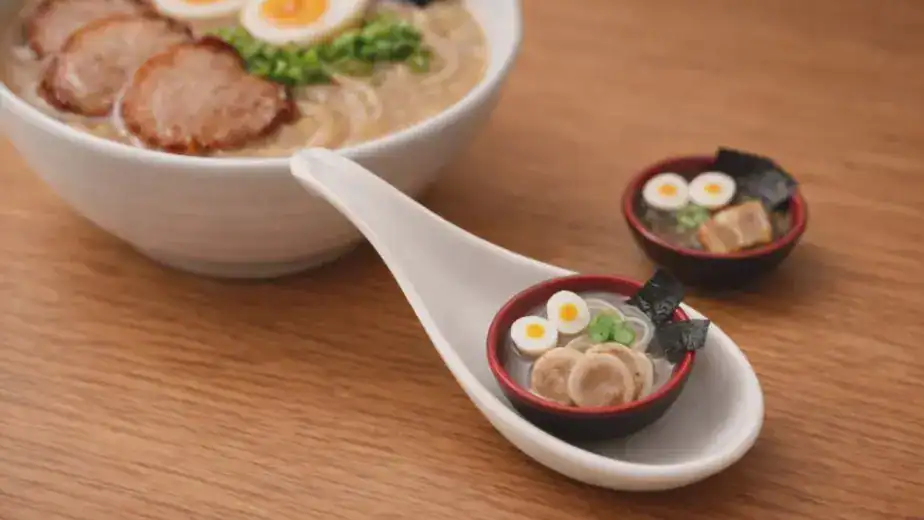 A tiny handcrafted miniature ramen bowl on a spoon, displayed alongside a full-size tonkotsu ramen bowl — made in RESOBOX's miniature food art class in New York