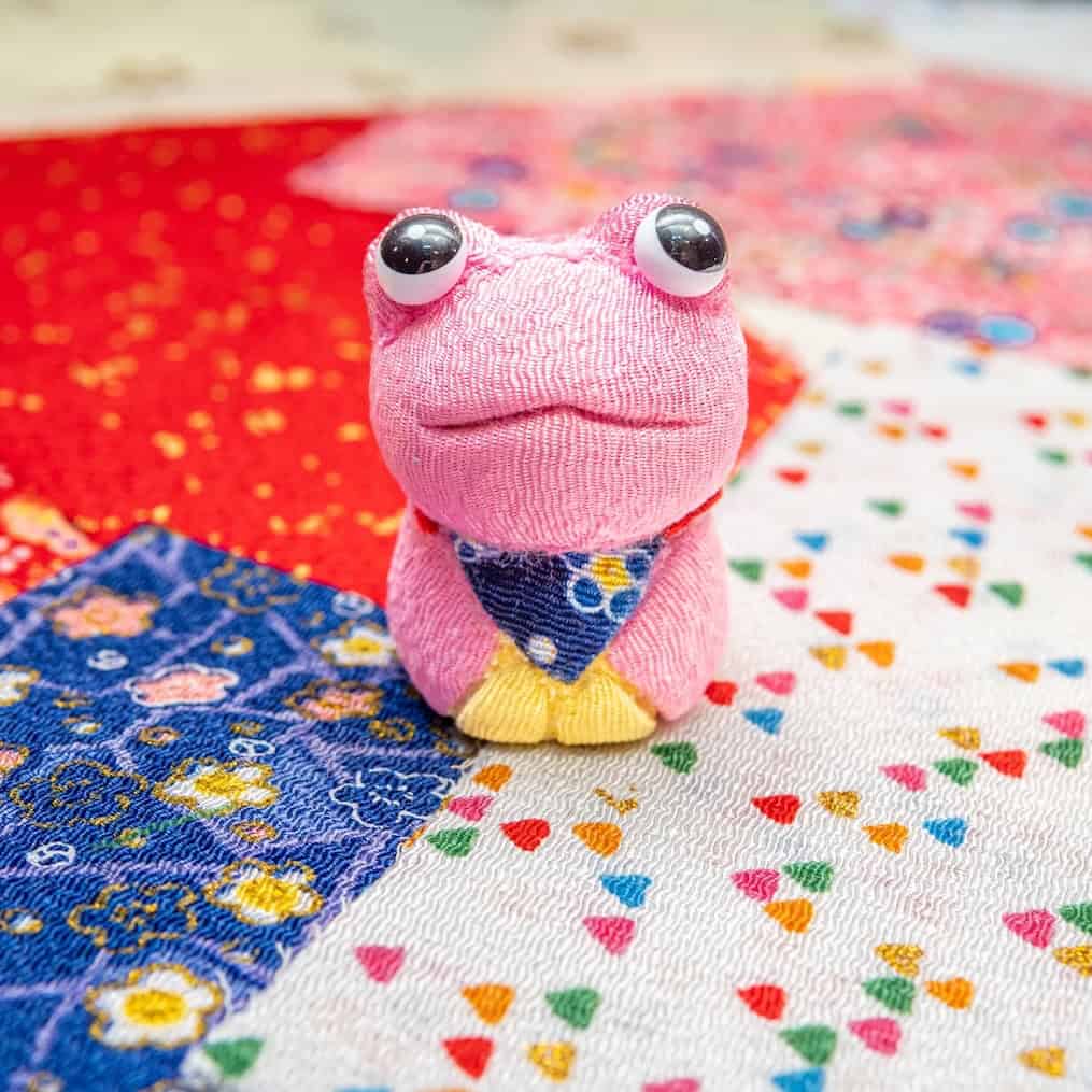 A handcrafted pink kimekomi frog doll with a navy fabric bib, displayed on colorful Japanese patterned fabrics — made in RESOBOX's kimekomi doll-making class in New York