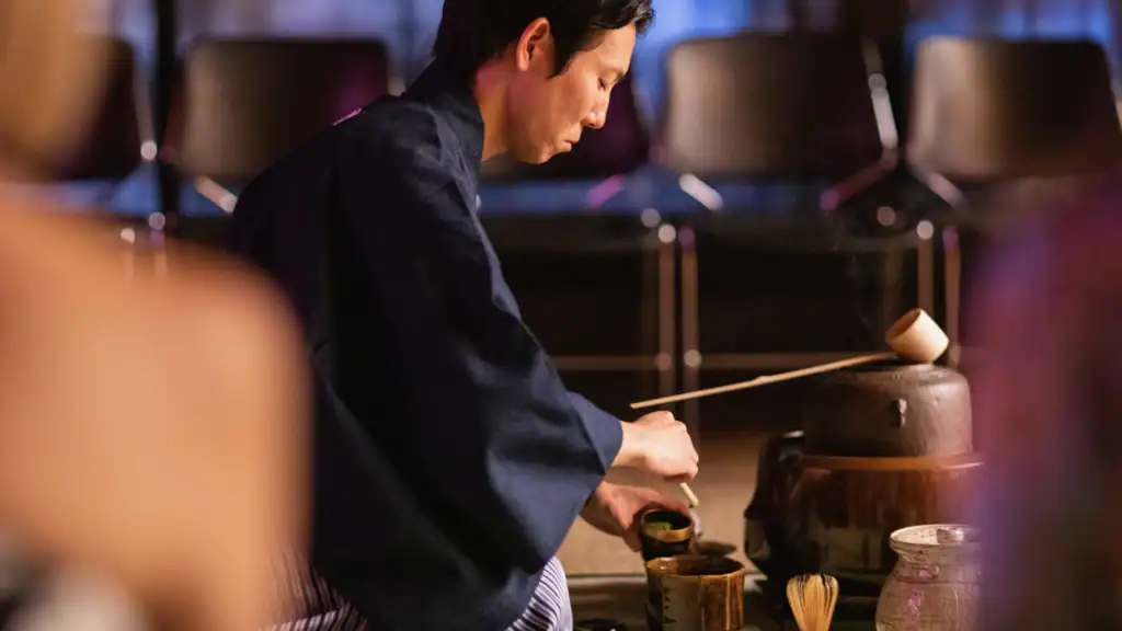 A tea master in a traditional navy kimono performing a formal Japanese tea ceremony demonstration, whisking matcha with a chasen in a dark ceramic bowl at RESOBOX in New York