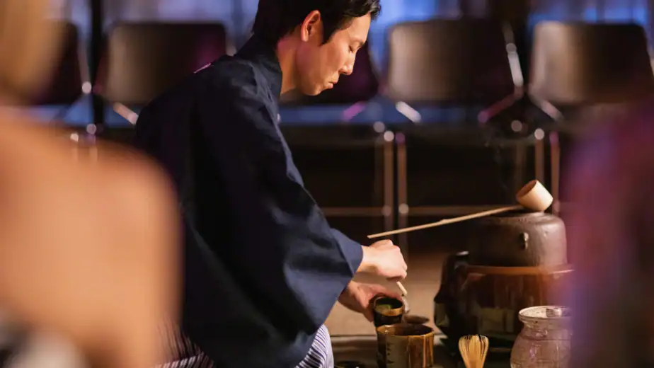 A tea master in a traditional navy kimono performing a formal Japanese tea ceremony demonstration, whisking matcha with a chasen in a dark ceramic bowl at RESOBOX in New York