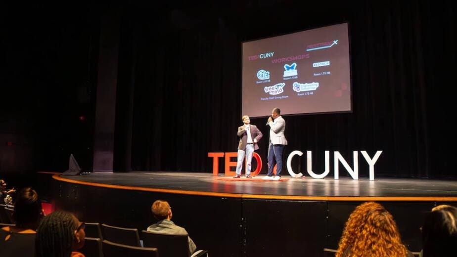 Two speakers presenting on TEDxCUNY stage with workshop logos displayed on projection screen, featuring Japanese cultural education programs and partner organizations in modern auditorium setting