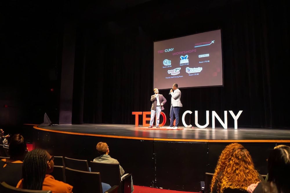 Two speakers presenting on TEDxCUNY stage with workshop logos displayed on projection screen, featuring Japanese cultural education programs and partner organizations in modern auditorium setting