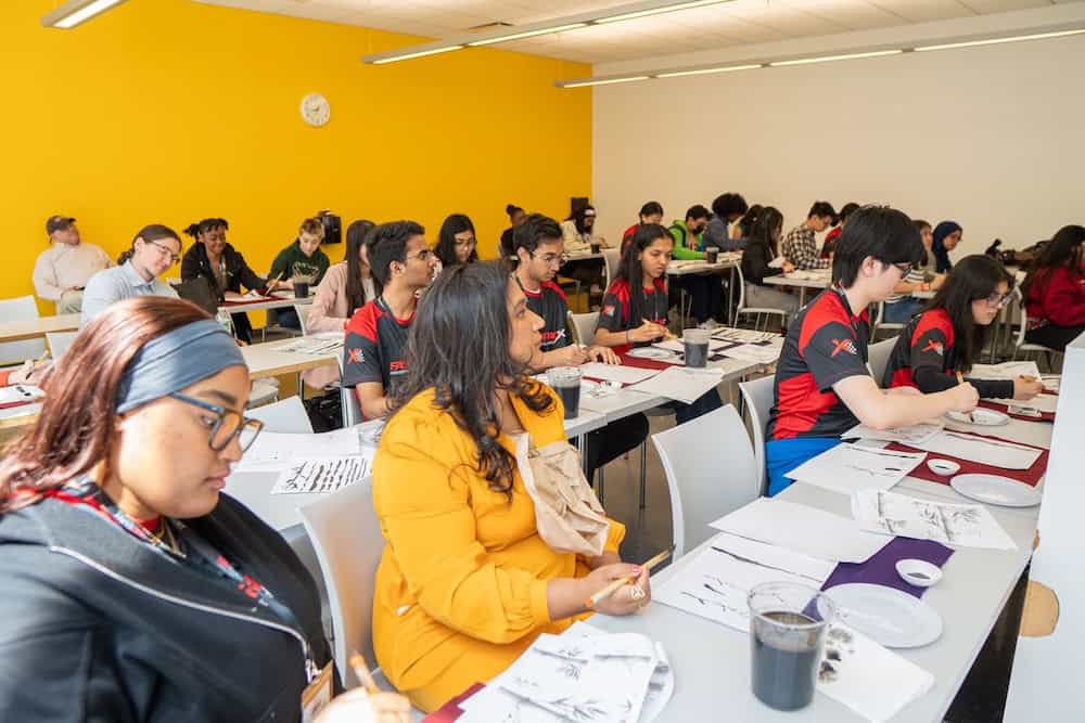 Large group of diverse participants attending sumi-e (Japanese ink painting) workshop in bright yellow-walled classroom at TEDxCUNY, with students engaged in traditional brush painting instruction