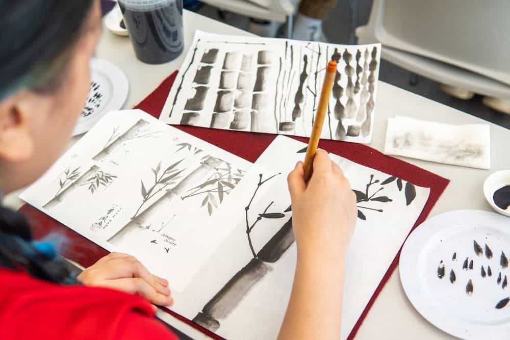 Close-up detail of participant practicing traditional sumi-e brush painting technique with Japanese ink and botanical motifs on white paper, demonstrating classical ink painting methods during TEDxCUNY workshop