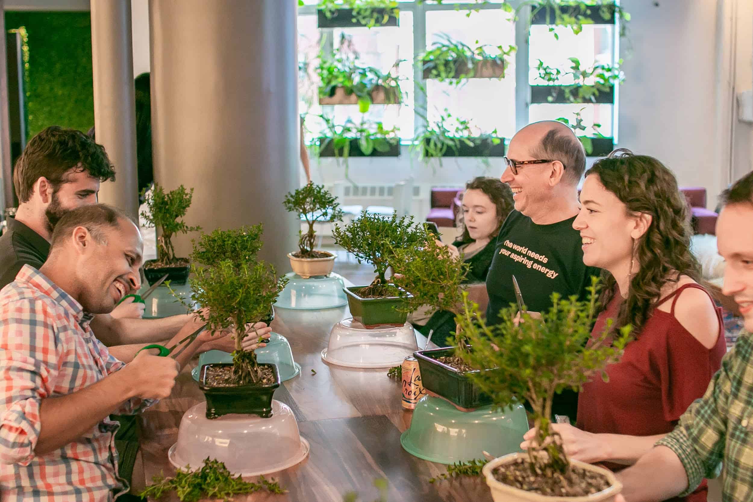 A group of smiling participants pruning their own bonsai trees around a long table during a bonsai workshop at RESOBOX in New York