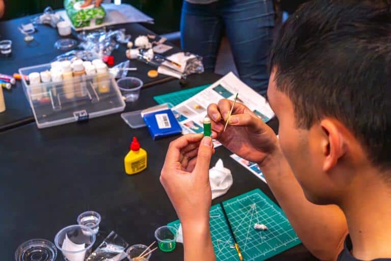 A participant painting a tiny miniature food piece with a fine brush, surrounded by paints and craft supplies on a workshop table