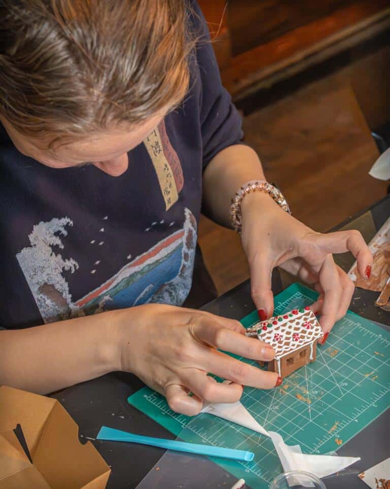 A participant carefully assembling a tiny decorated gingerbread house with red and green icing at a hands-on workshop