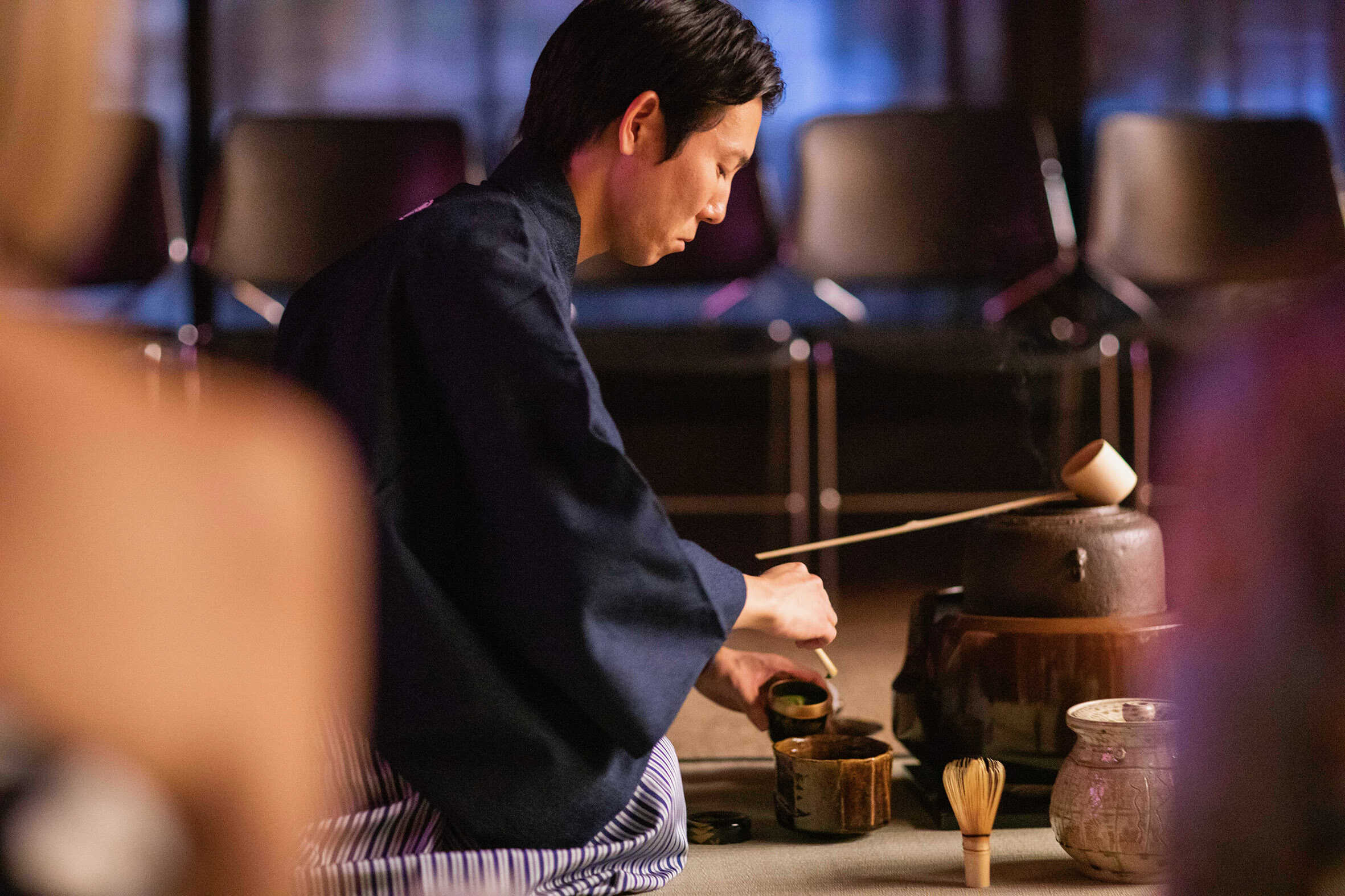 Tea master Yoshitsugu Nagano in kimono performing a traditional chanoyu tea ceremony, whisking matcha beside a ceramic water jar before an audience