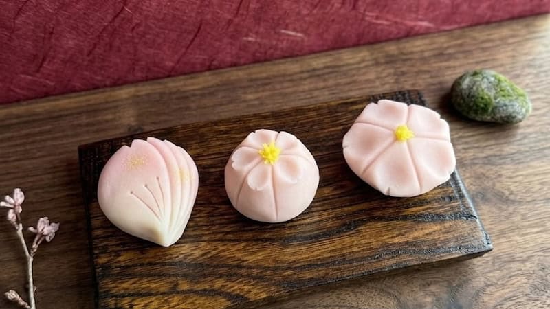 Three handcrafted pink wagashi sweets shaped as a cherry blossom petal, chrysanthemum, and morning glory, displayed on a wooden board during a Japanese sweets class at RESOBOX in New York