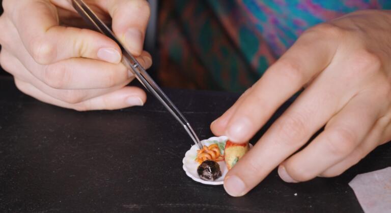 A participant carefully placing miniature food pieces onto a tiny plate using tweezers during a RESOBOX miniature food-making class