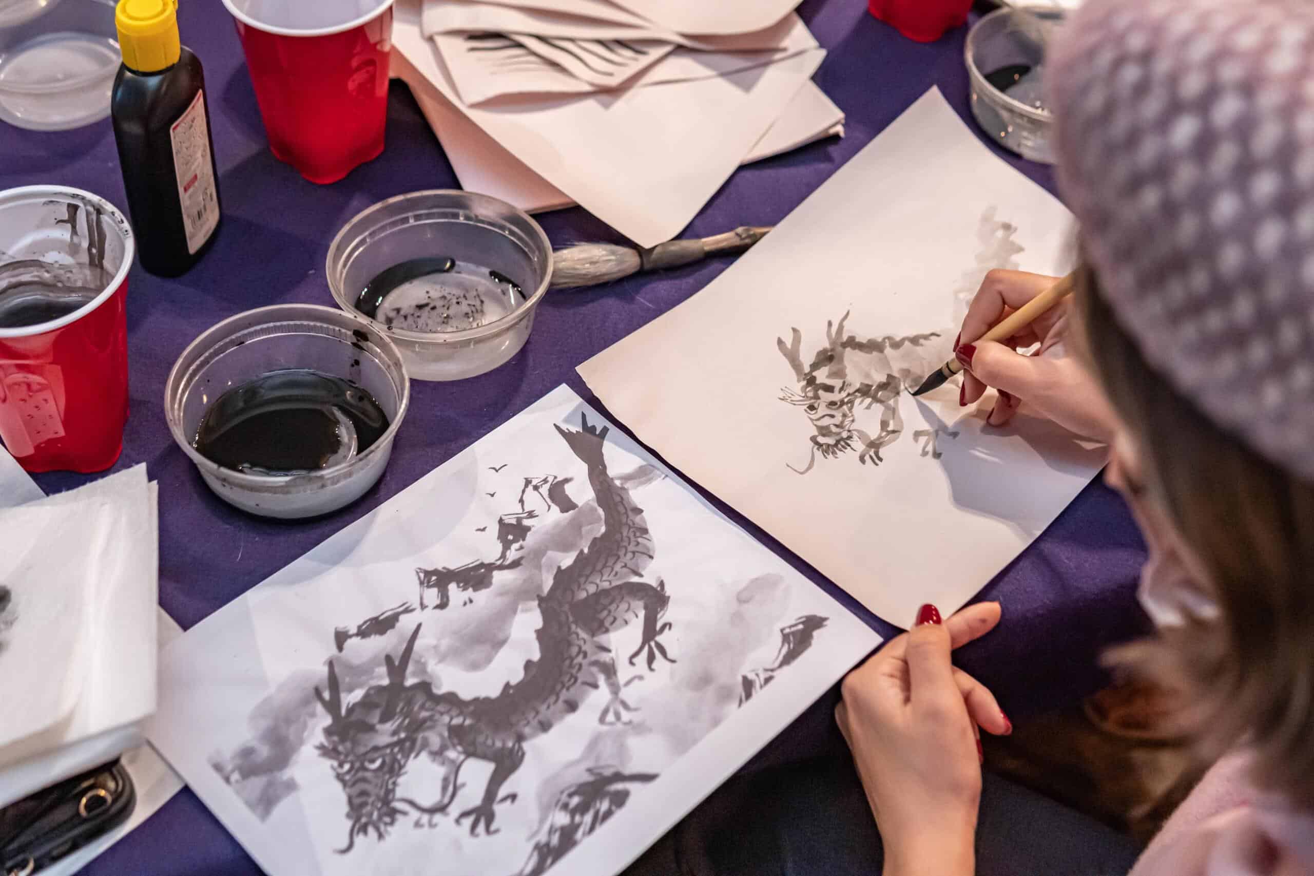 A participant painting a dragon in Japanese sumi-e ink wash style, referencing a detailed example sheet, during a sumi-e class at RESOBOX in New York