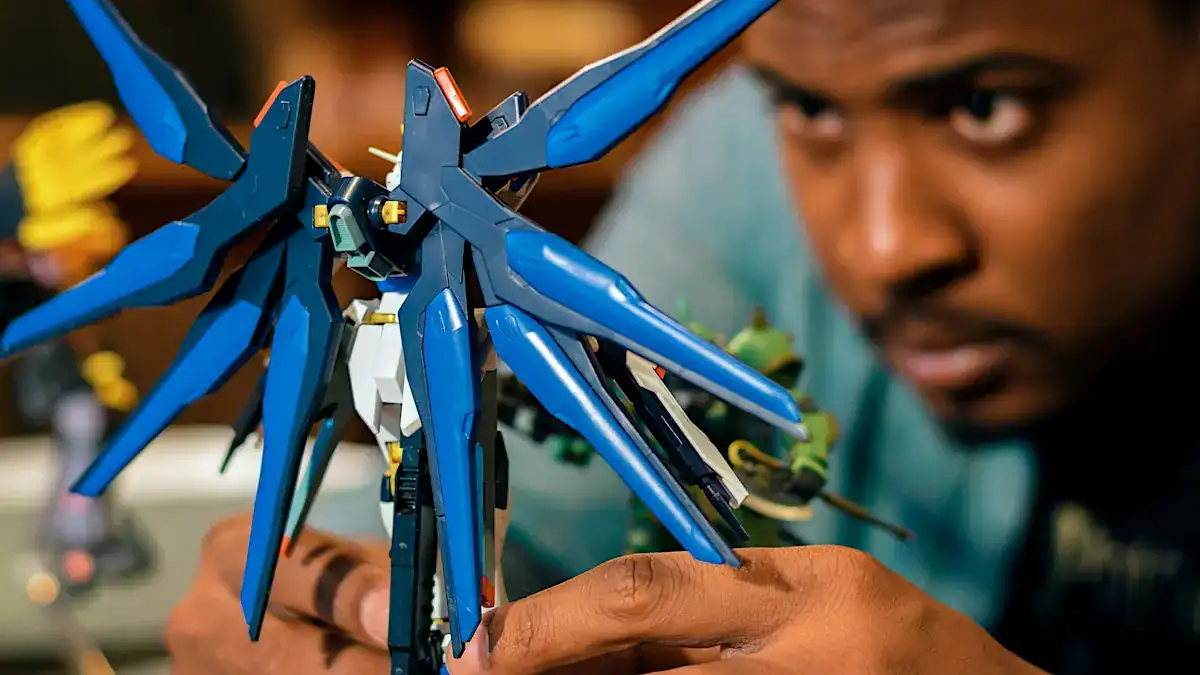 Tim Martin-Toussaint closely inspecting a completed blue Gundam model kit with large wing binders at a Gunpla build night