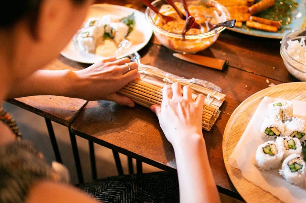 A participant rolling sushi using a bamboo mat during a hands-on sushi making class at RESOBOX in New York, with freshly made California rolls and ingredients spread across a wooden table