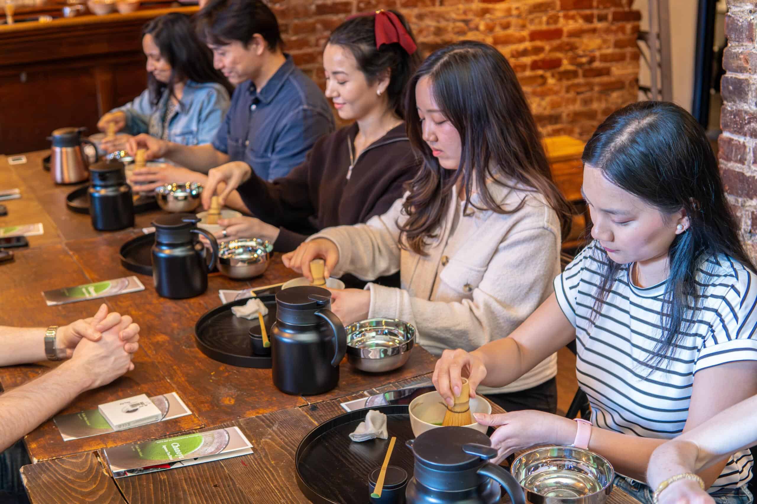 Participants whisking matcha in traditional bowls during a Japanese tea ceremony class at RESOBOX in New York's East Village, seated along a wooden table in a warm brick-walled studio