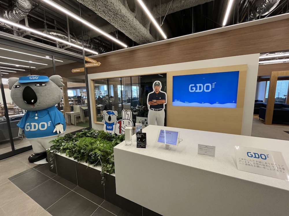 GDO(ゴルフ用品大手)のオフィス内に展示された、様々なゴルフ関連製品と情報パネルが配置された木製棚。プロゴルファーやアマチュア向けのスポーツシューズやアクセサリーが整理されている