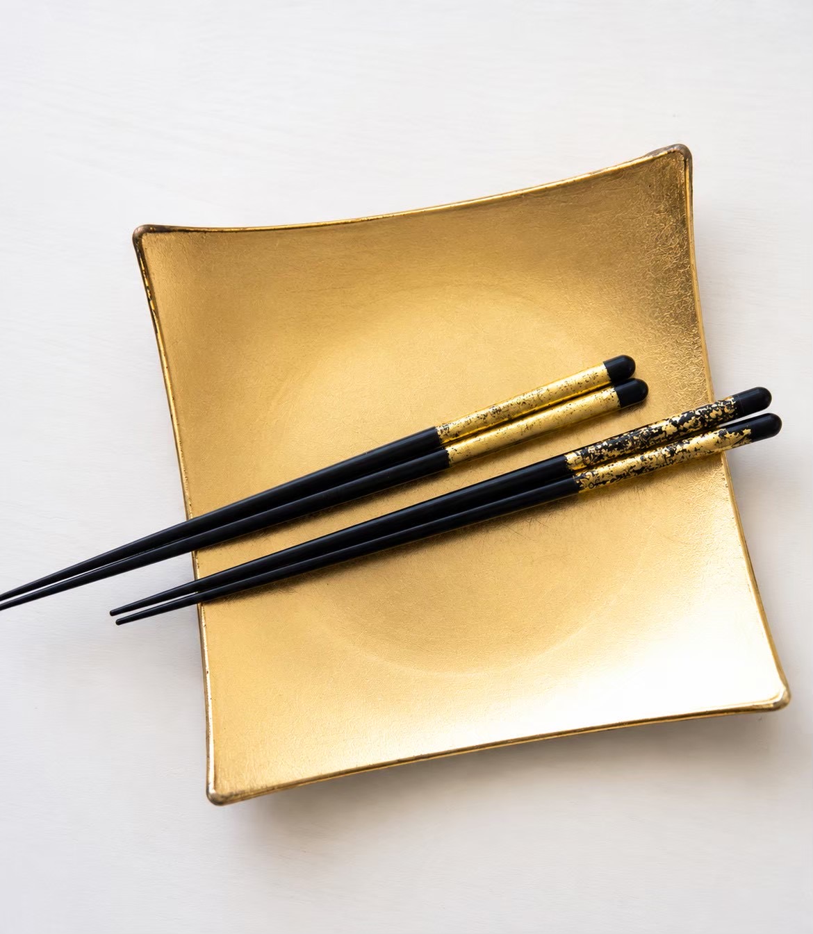 Two pairs of chopsticks with different gold leaf designs laid across a square gold-leaf dish