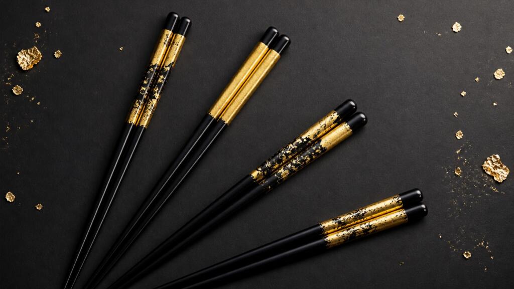 Four pairs of black chopsticks decorated with Kanazawa gold leaf in four different designs, on a dark background scattered with gold flakes