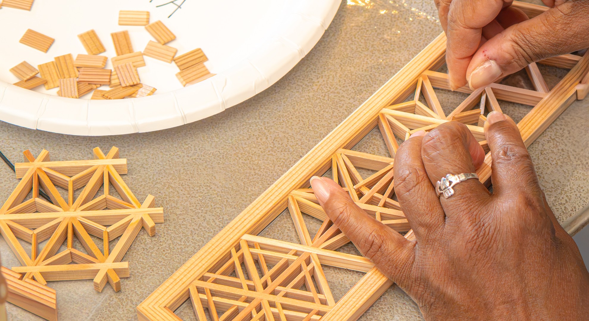 Close-up of hands assembling an asa-no-ha kumiko pattern beside a finished lattice piece at a kumiko woodworking class at RESOBOX in New York City