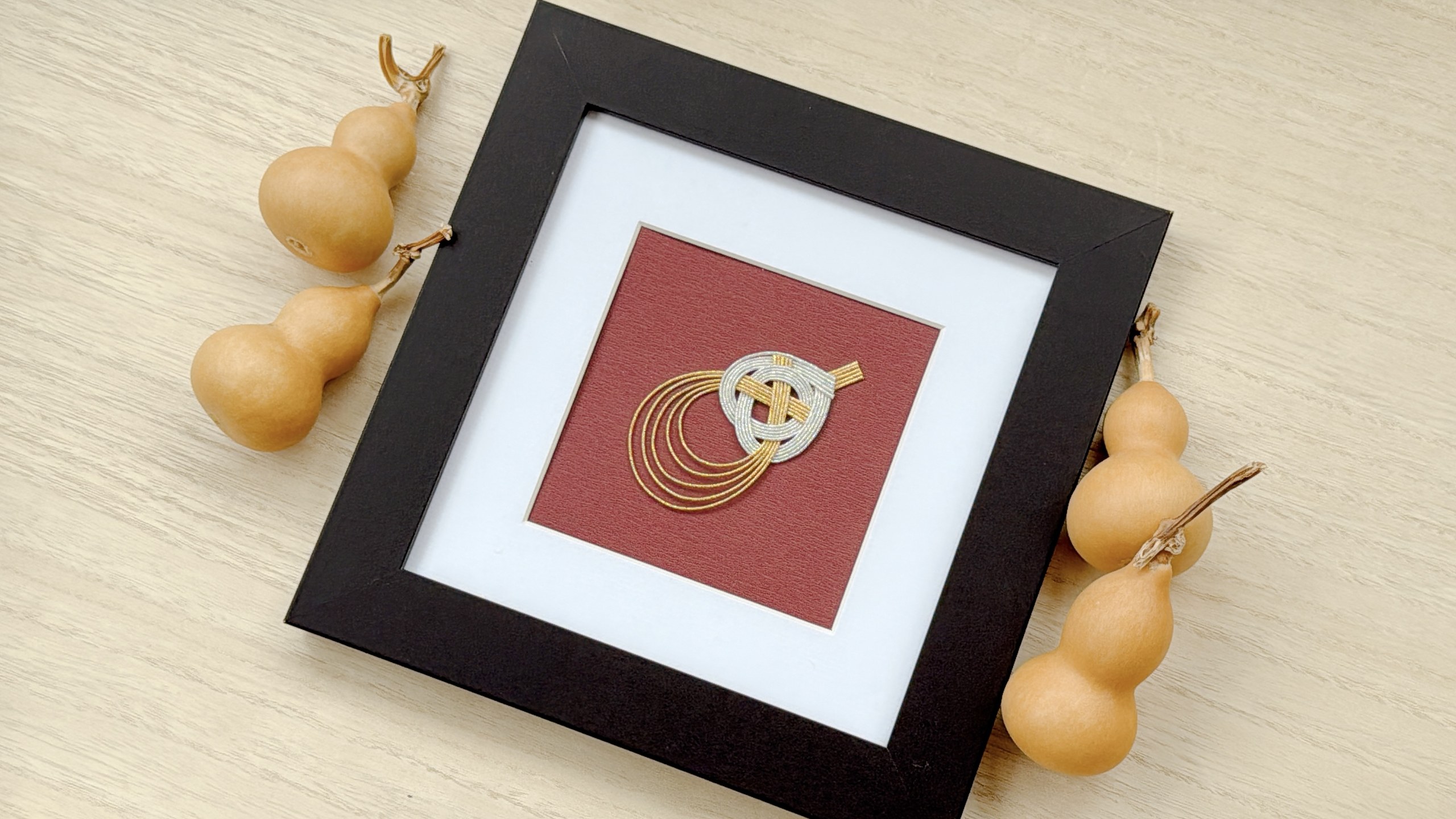 Framed mizuhiki gourd artwork in gold and silver cord, displayed beside dried gourds