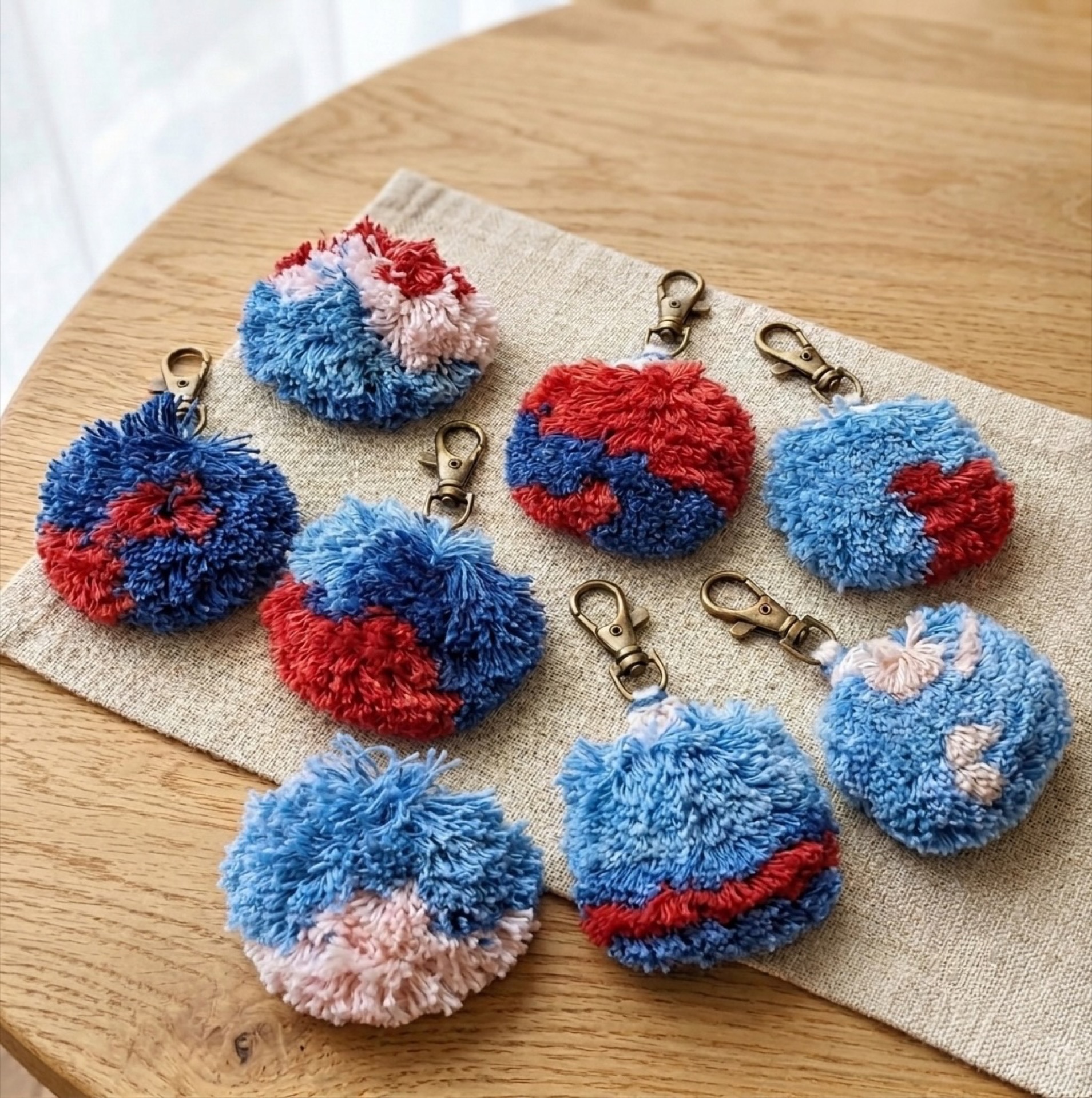 Nine finished hand-knotted pieces in indigo, red and pale pink, each on a brass keyring clip, laid out on linen.