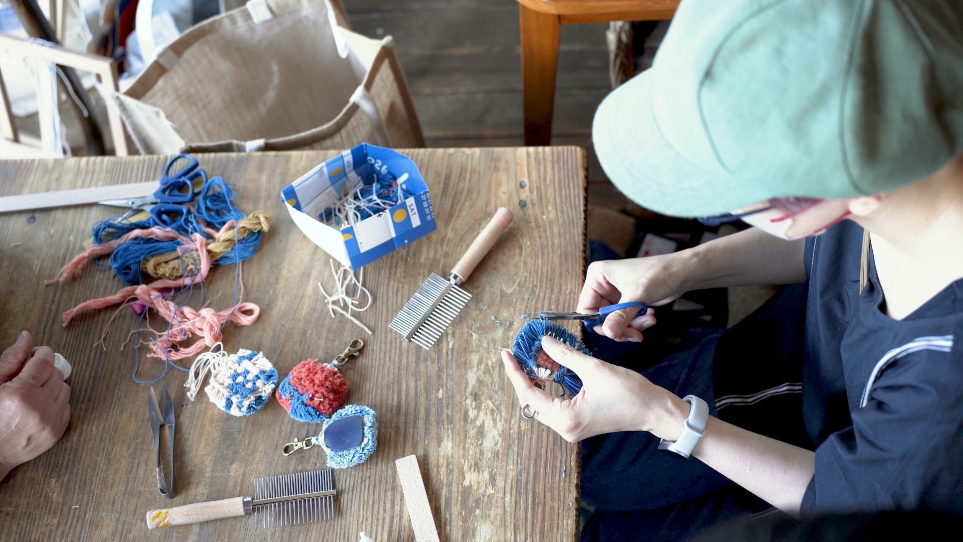 A worktable seen from above during a workshop in Japan: plant-dyed cotton in blue and coral, two steel combs, a pair of Japanese hand shears and several finished knotted charms.