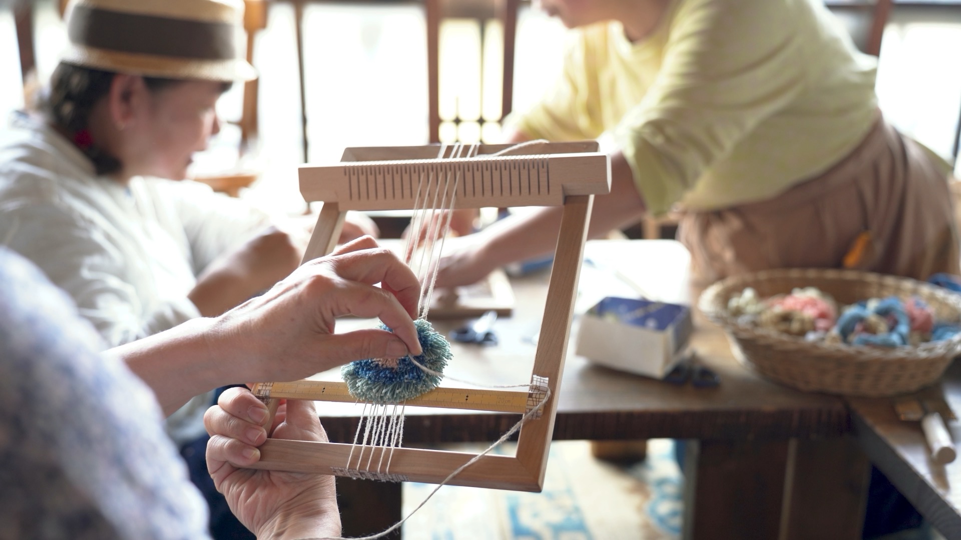 Hands holding a small wooden frame loom, tying a tuft of blue dyed cotton onto the warp threads.