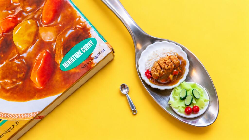 A miniature Japanese katsu curry plate and a small side salad on tiny scalloped dishes, resting on a full-size spoon next to a tiny spoon