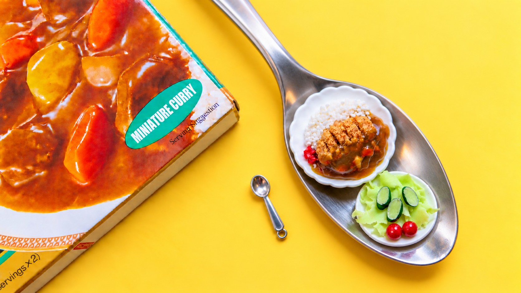 A miniature Japanese katsu curry plate and a small side salad on tiny scalloped dishes, resting on a full-size spoon next to a tiny spoon