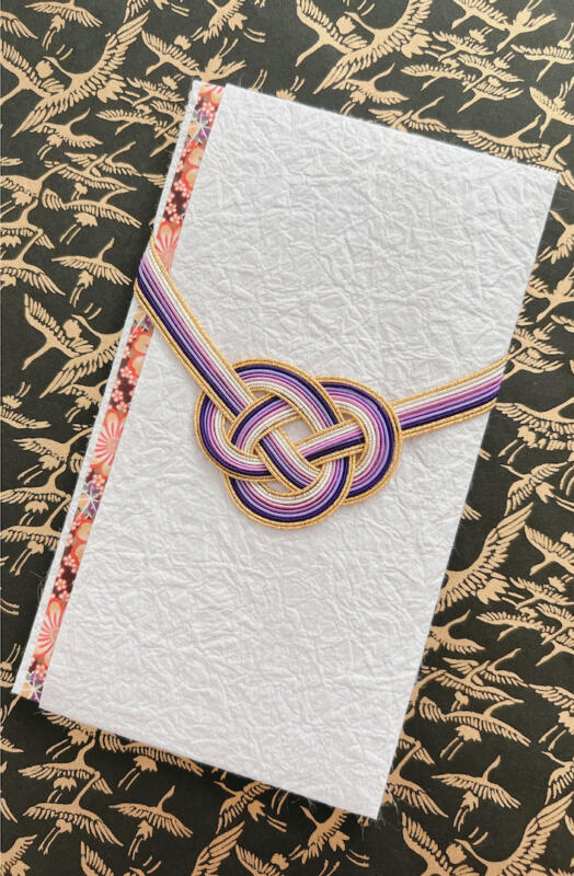 A traditional Japanese washi envelope adorned with a handcrafted purple and gold mizuhiki awaji knot, displayed on a crane-patterned background — made in RESOBOX's mizuhiki class in New York