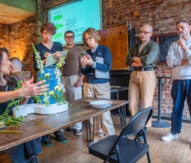 An ikebana instructor explaining flower arrangement techniques to an attentive group of students gathered around a display at RESOBOX