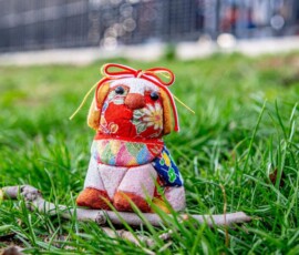 A handmade kimekomi dog doll crafted with colorful patterned chirimen fabric and a red ribbon, displayed on green grass outdoors