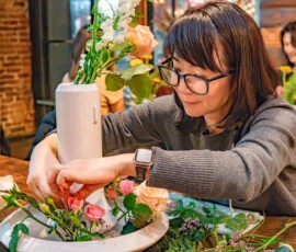 A participant carefully placing pink and peach roses around a tall white vase during a moribana ikebana flower arrangement class at RESOBOX