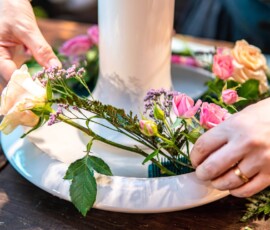 Close-up of hands arranging pink and peach roses with statice around a kenzan in a white suiban dish during an ikebana flower arrangement class