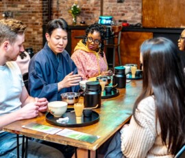A kimono-clad tea ceremony instructor explaining the art of chanoyu to an attentive group of participants at RESOBOX, with matcha tools arranged on black trays