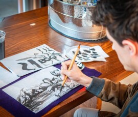 A participant adding detailed brushwork to a sumi-e ink painting of a mountain landscape with flowers, with ink cup and practice sheets nearby