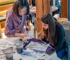 A sumi-e instructor guiding a student's brushwork on a cherry blossom ink painting, with ink dishes and practice sheets spread across the table