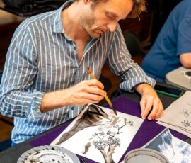 A participant applying ink brushstrokes to a sumi-e painting of a cherry blossom tree during a Japanese ink wash painting class
