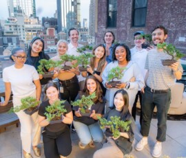 A group of participants proudly displaying their pruned juniper bonsai trees after a rooftop bonsai class, with the New York City skyline in the background