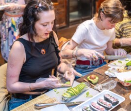 A participant carefully pressing sliced avocado onto a sushi roll during a kazari maki zushi workshop, with completed decorative rolls on a white plate in the foreground