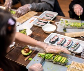 Participants layering sliced avocado over sushi rolls on a cutting board during a kazari maki zushi workshop, with completed decorative rolls on a white plate nearby