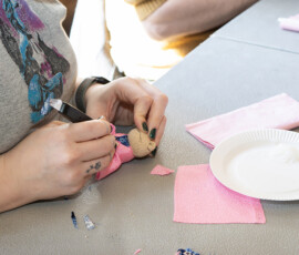 Hands carefully tucking pink fabric onto a small kimekomi doll base with a tucking tool, with glue and fabric scraps on the worktable