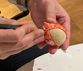 Hands tucking red chirimen fabric into the grooves of a kimekomi doll base using a tucking tool during a kimekomi doll-making class